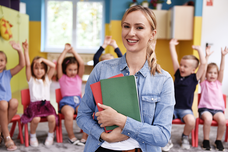 Educator smiling and holding folders with seated children in the background